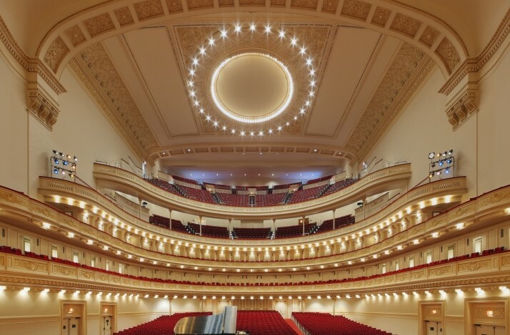 A grand, ornate concert hall interior of Carnegie Hall, surrounded by multiple tiers of red seating.