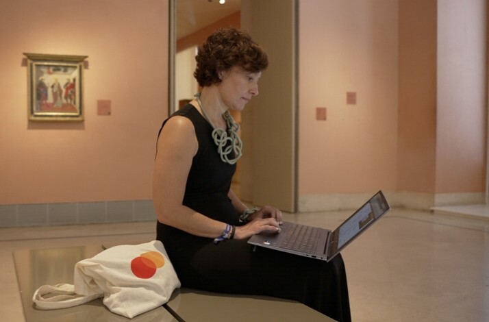 Rocío Rubio Álvare seated and engrossed in her laptop, with a Mastercard logo tote bag rests beside her on the bench. 