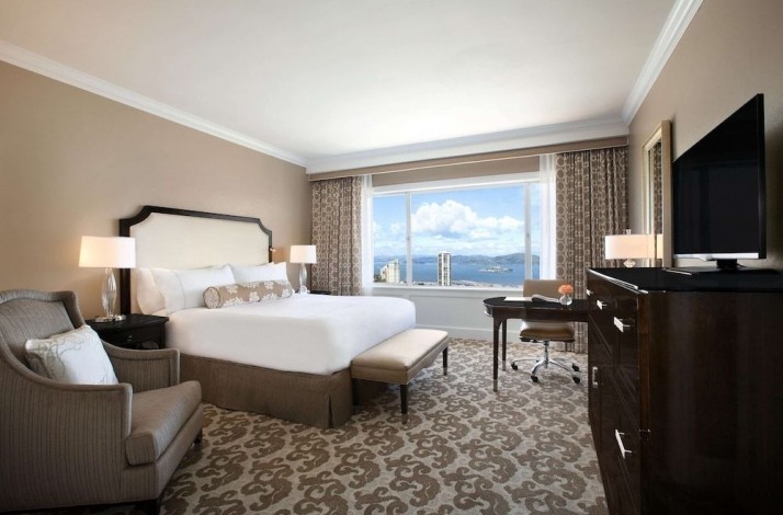 A bedroom at one of the Fairmont San Francisco hotel rooms. The room is furnished in beige colors and has a panoramic window.