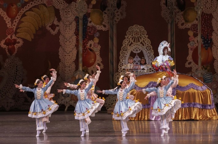 Dancers in pale blue costumes perform in formation in front of a large gilded bed on an elaborate set.