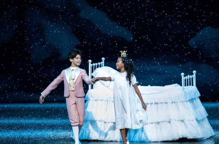 A boy in a pink suit holds hands with a girl in a white nightgown beside a large bed as snow falls onstage.