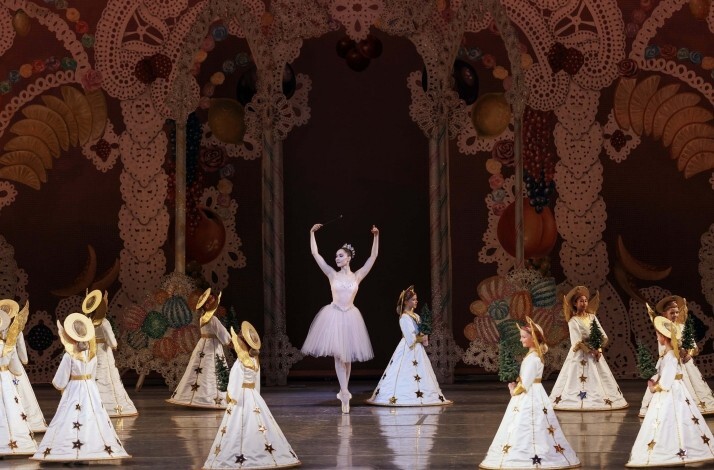 A ballerina in a white tutu stands en pointe center stage as children in white angel costumes circle around her.