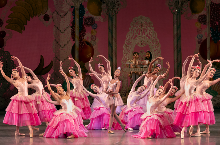 Ballet dancers in pink dresses pose in a semicircle onstage around a central ballerina, with a bright pink, candy-themed backdrop.