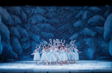 A group of ballet dancers in white tutus pose with raised arms as artificial snow falls on a blue, ice-like stage set.