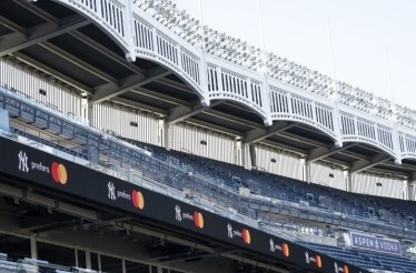 A view of a baseball stadium’s upper deck with empty seats and bright light rigs, featuring a ribbon board repeating NY prefers and an ASPEN VODKA sign.