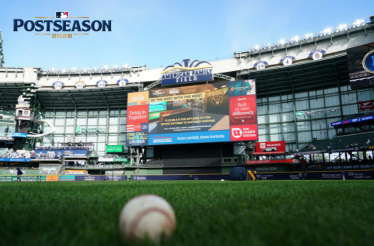 Baseball resting on American Family Field grass, with the stadium scoreboard and MLB Postseason 2026 logo visible in upper-left corner.