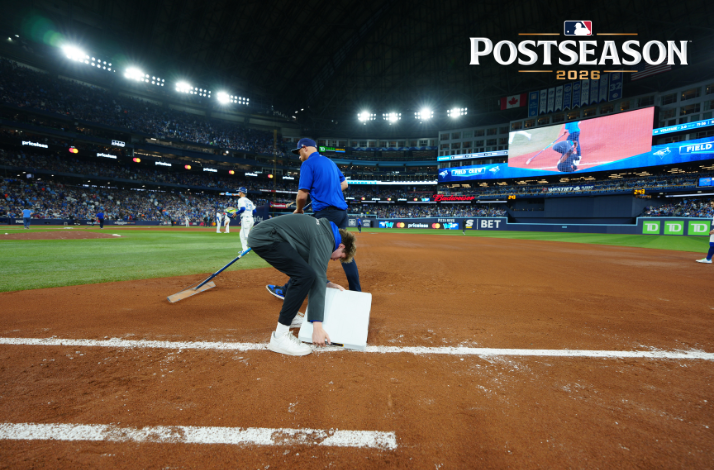 Grounds crew preparing the field at a packed MLB Postseason 2026 stadium, with a large scoreboard visible. Postseason 2026 logo in the top right corner.