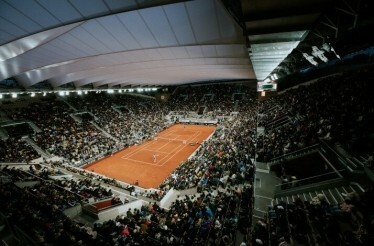 A fan's wide view of a live tennis game in Roland-Garros stadium, filled with fans and an open telescopic roof.