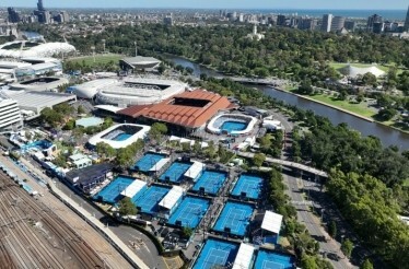 Outdoor tennis complex with multiple blue courts, stadiums, and surrounding cityscape.