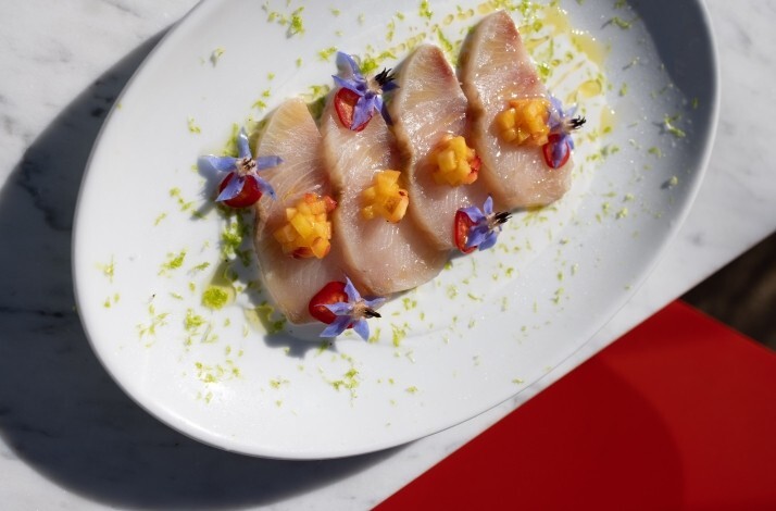 Elegant crudo course garnished with purple edible flowers, diced fruit, and citrus zest, showcasing Chef Levi's seasonal approach.