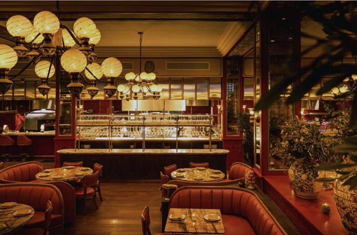 Warmly lit dining room at Mother Wolf LA, featuring red leather banquettes, marble bar, globe chandeliers, and an open kitchen view.