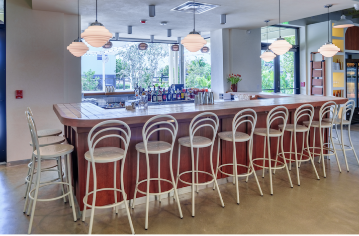 Spacious bar at Bar Bucce Miami featuring a wood-topped counter, white bistro stools, globe pendant lights, and an open-air view.