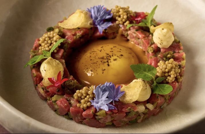 Beef tartare ring centered with egg yolk, garnished with blue cornflowers, mustard seeds, crisps, and fresh herbs at MILA Miami Beach.