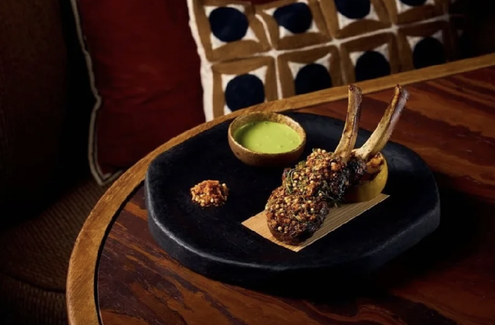 Fire-cooked lamb chops coated in toasted seeds and herbs, plated on a dark slate board alongside a vibrant green sauce.