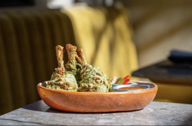 Three green herb-coated bone-in meat pieces arranged in a terracotta bowl alongside a small sauce vessel at Si! Mon, Venice.
