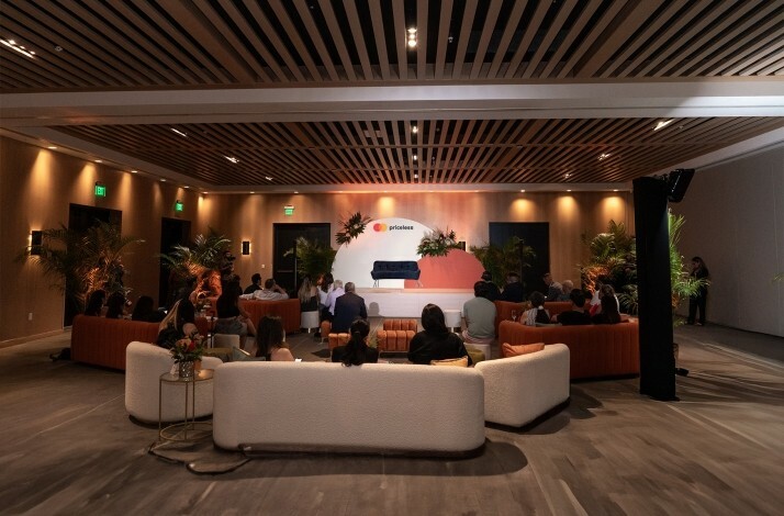 A large room with guests seating in front of a small stage with priceless branding on the background.