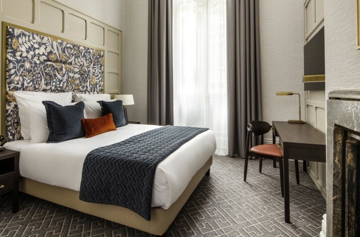 Modern hotel room with a double bed, patterned headboard, desk, and large window with curtains