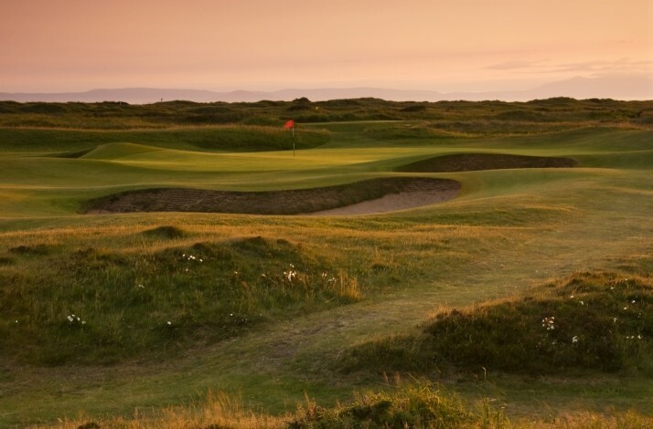 Golf course at sunset with a red flag and sand bunker on rolling green fairways.