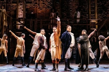 The cast of Hamilton performing a dynamic scene on stage, featuring period costumes and choreography.
