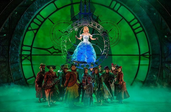 Actors on stage during a play, a blue gown wearing female actor standing above the others inside the mechanics of a huge clock.