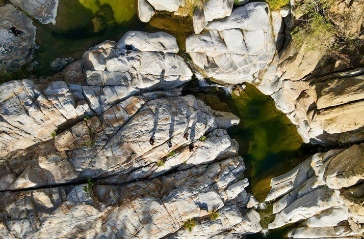 Aerial view of rocky formations and green pools at Cañón de la Zorra in Mexico's Sierra de la Laguna, with people visible on the rocks.