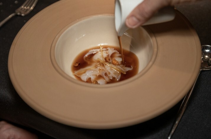 A hand pours dark brown sauce over delicate white dumplings garnished with thin strips and crumbs in a beige bowl, creating a warm, enticing scene.