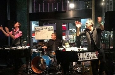 Two of the top piano entertainers in NYC playing songs on stage joined by a drummer.