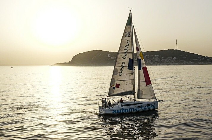 A sailboat with logos on its sails cruising on the sea during a golden sunset, with an island silhouette and sun reflections on the water in the background.