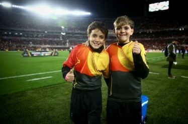 Two young boys, smiling and giving thumbs up, stand on a football pitch stadium during a game, wearing team jerseys.
