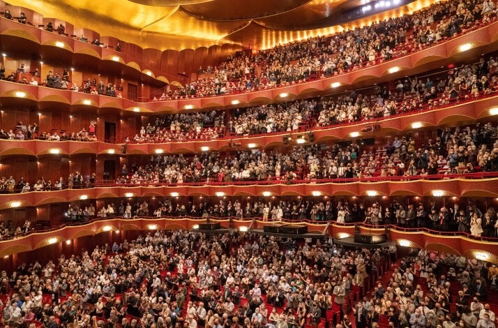 Packed The Metropolitan Opera interior with multiple red-and-gold balconies and a standing audience applauding.
