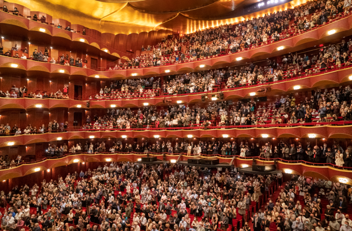 The image shows the interior of the Metropolitan Opera House in New York City, which is part of Lincoln Center for the Performing Arts with a lot of persons