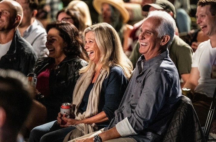 A diverse audience enjoys a performance, with some attendees holding drinks and wearing casual attire.