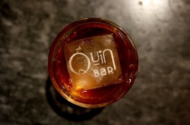 A crafted drink garnished with the Quin Bar logo.
