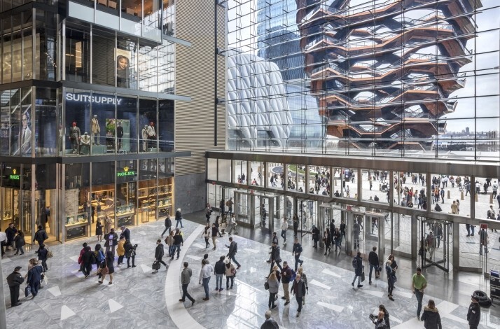 A bustling retail concourse showcasing storefronts and the Vessel through glass walls.