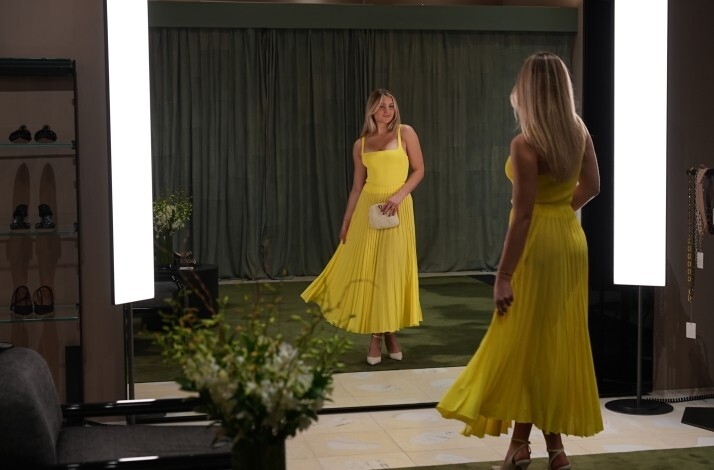 A lady admiring a bright yellow dress in an elegant fitting room with large mirrors.