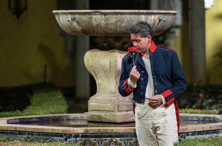 Performer in 19th-century military costume smelling a red rose beside a stone fountain at Palacio de Las Dueñas.