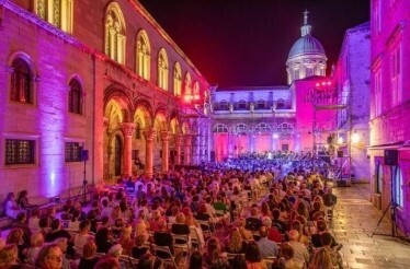 An open-air classical concert at night in Dubrovnik Summer Festival, with historic buildings illuminated in bright pink light.