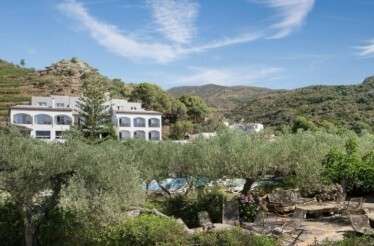 Hotel Restaurante Cala Joncols' white multi-storey building nestled among olive trees, gardens, and terraced hillside.