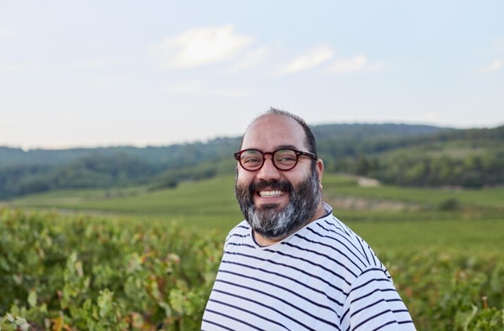 A smiling man wearing glasses standing in a vineyard with rolling green hills in the background.