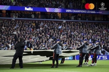 Staff unfolding a pitch cover across a stadium, Mastercard and UEFA Champions League 'official sponsor' logos in the upper-right corner.