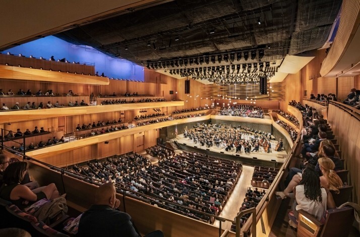 Wide view of the New York Philharmonic hall filled with audience members, with an orchestra performing onstage.