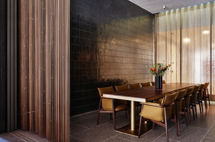 A dining area features a wooden table with mustard-colored chairs, set against a wall of dark-brown, rectangular tiles. 