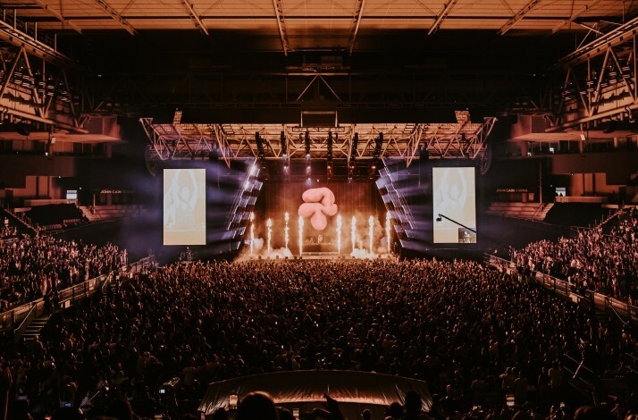 Packed crowd at John Cain Arena during AO Live Presents, with pyrotechnics, beam lighting, and a large inflatable prop illuminating the stage.
