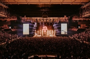 Packed crowd at John Cain Arena during AO Live Presents, with pyrotechnics, beam lighting, and a large inflatable prop illuminating the stage.