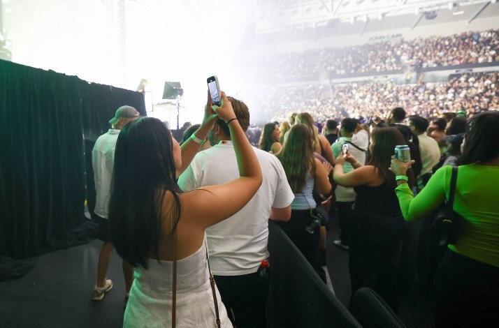 Side-of-stage view at John Cain Arena, with a Mastercard cardholder filming AO Live Presents while crowds fill the arena in the background.