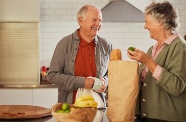 An old-couple smiling each other with a groceries bag in a table  