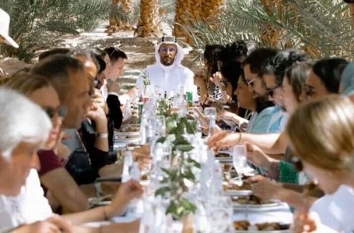 A long table set in the desert with a group of people gathered around, enjoying a meal in a serene outdoor setting.