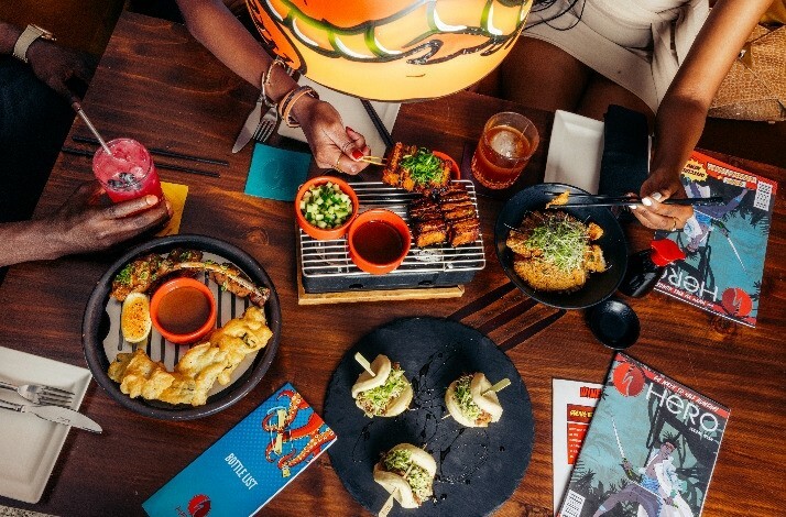 An overhead view of a vibrant dining table featuring a variety of dishes, including grilled meats, appetizers, and drinks, alongside magazines and a menu.