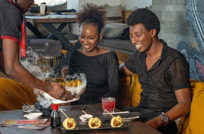 A server brings a steaming dish to a table where two diners sit, with colorful drinks and a plate of food in front of them.