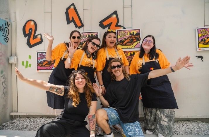 A group of six people smiling and posing together in front of colorful spray-paint artwork and graffiti-style numbers at an outdoor art workshop.